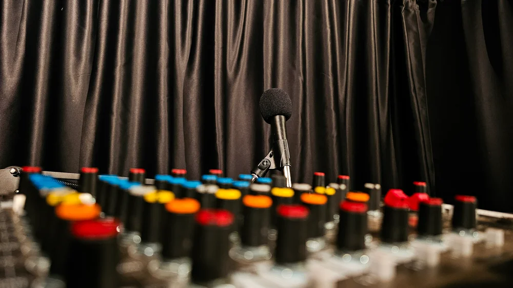 Tuvplāns ar audio mikšerpulti un tās krāsainajiem slēdžiem, fonā redzams mikrofons uz statīva pie tumšiem aizkariem.