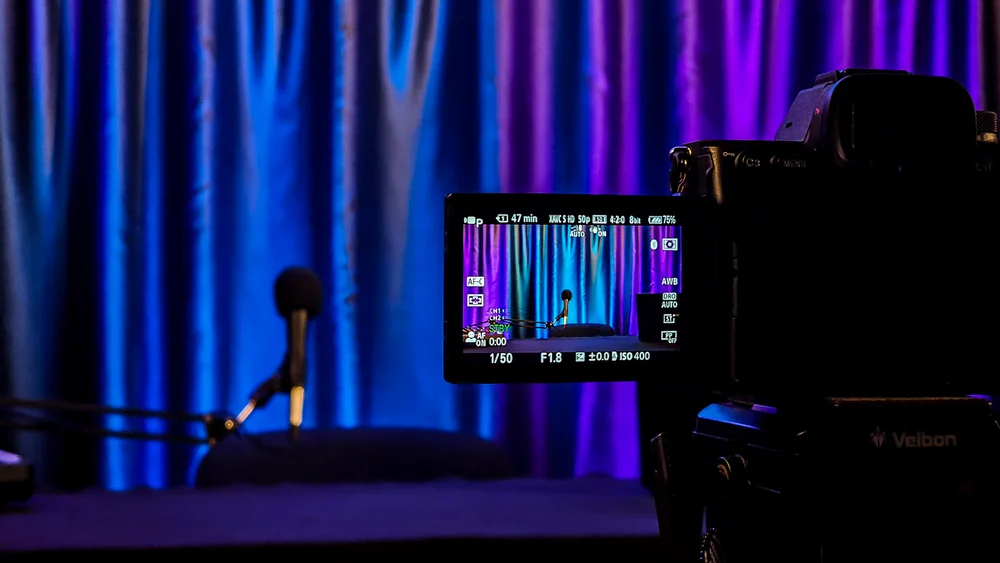 Tuvplāns ar video kameru, kas filmē podkāstu studiju ar violetu fona apgaismojumu.